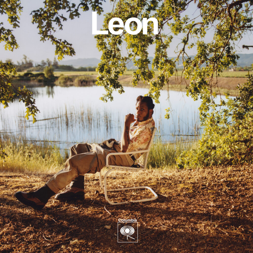 The image features singer Leon Bridges seated in a relaxed pose on a folding chair beside a serene lake, surrounded by grassy fields and trees under clear skies. He is wearing a casual, patterned short-sleeved shirt and light-colored pants, with one hand touching his chin thoughtfully. In the upper left corner, the text 