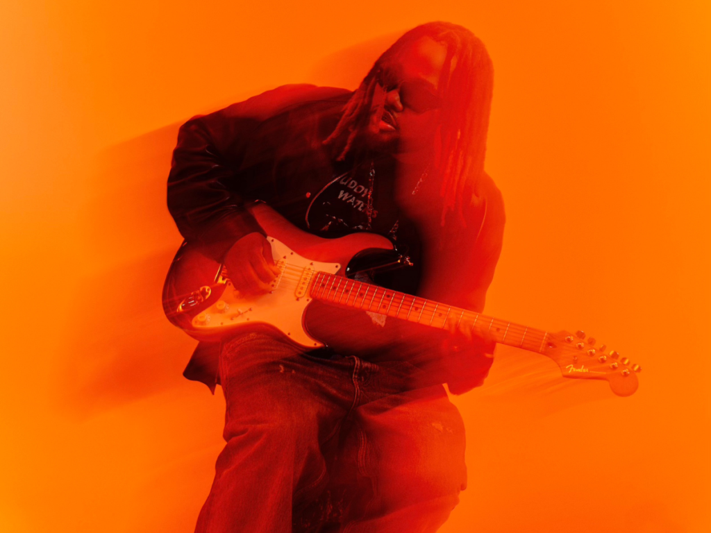 A photo of singer Leon Thomas holding an electric guitar against an orange backdrop